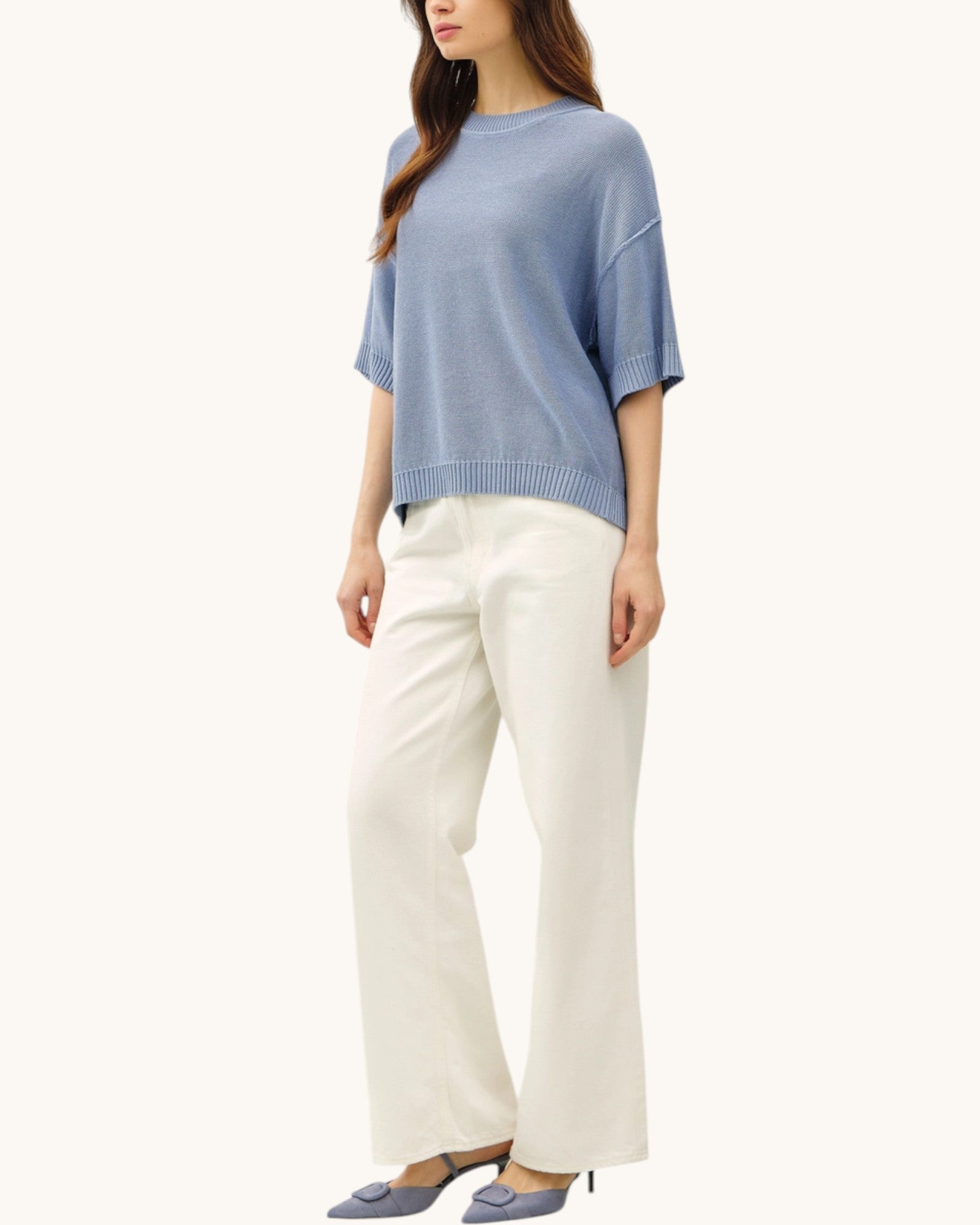 Woman wearing a blue lightweight sweater and white pants on a white background.