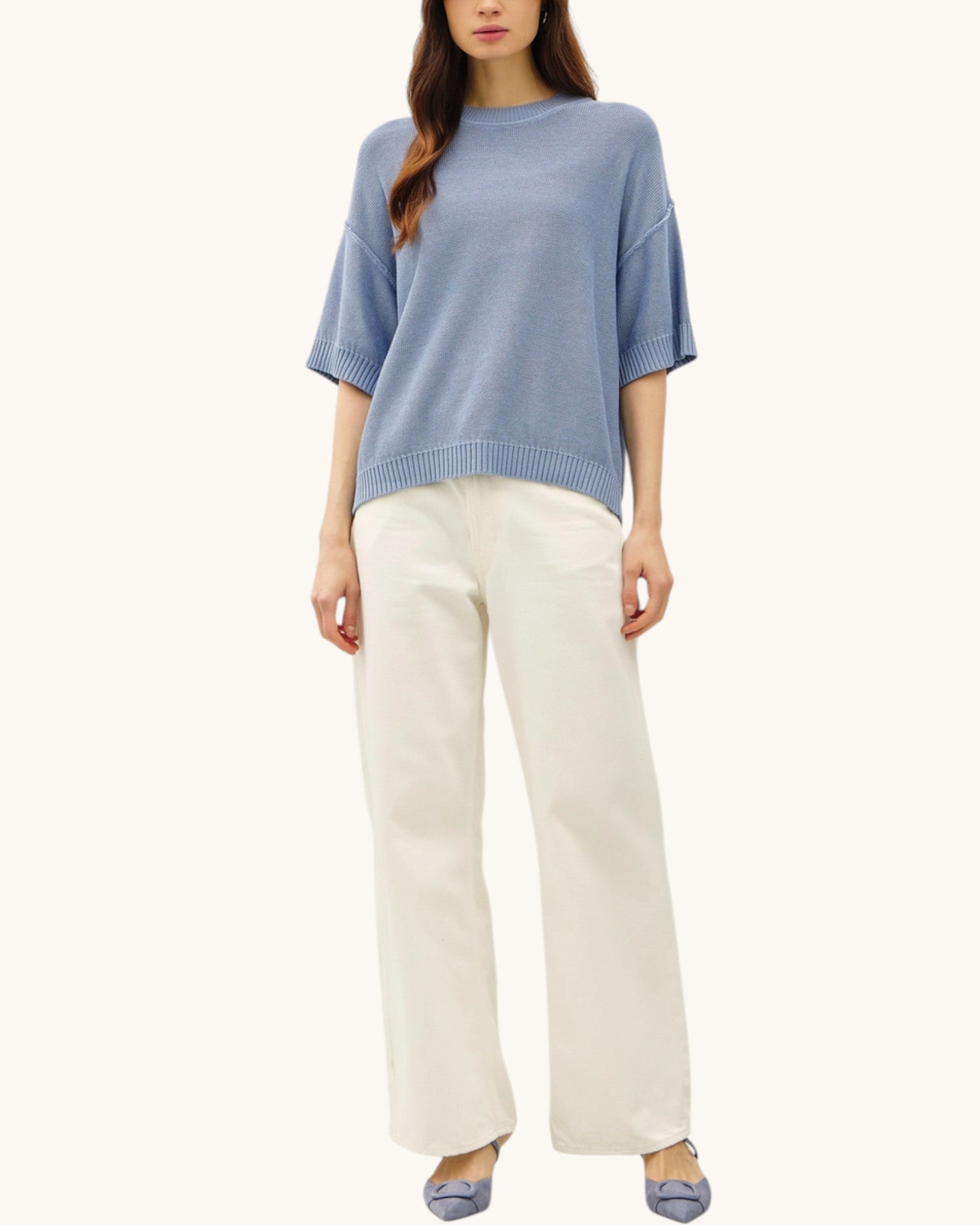 Woman wearing a short sleeve blue relaxed fit sweater and white pants on a white background.