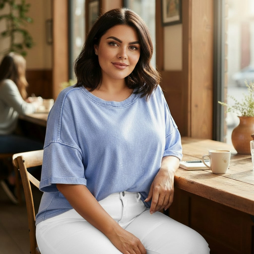 Woman sitting at a table in a cozy cafe wearing a blue shirt and white pants.