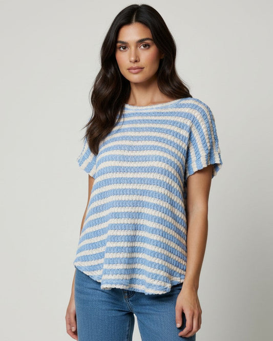 A woman modeling a short-sleeve knit top with horizontal light blue and off-white stripes, paired with blue jeans, against a plain light gray studio background.