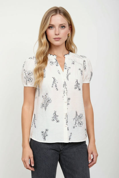 A woman modeling a white short-sleeve blouse with a black floral line-art print, paired with dark gray jeans, against a plain light gray studio background.