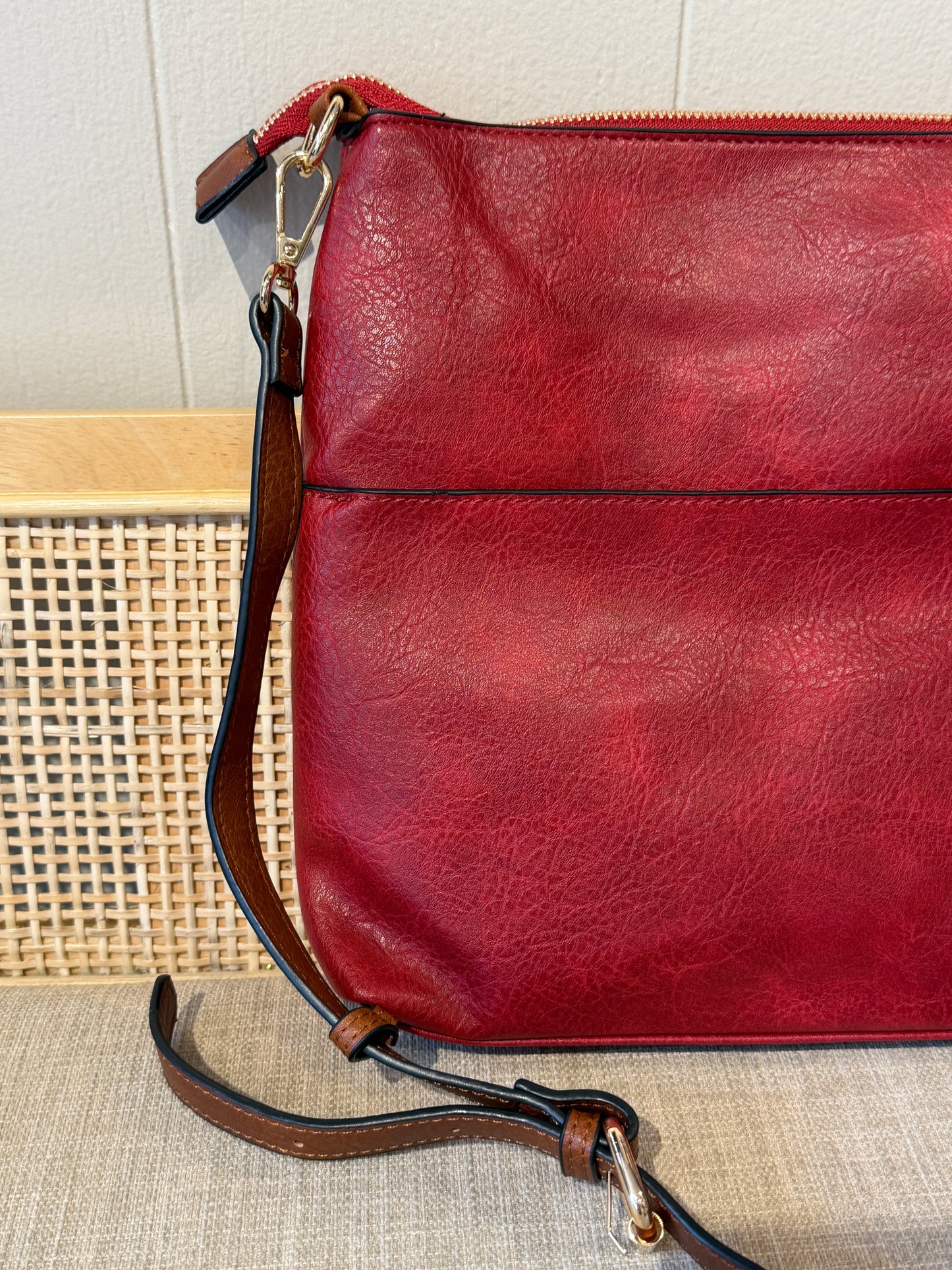 A close-up of a red leather crossbody bag with a brown leather strap and gold-tone hardware, resting on a woven surface against a light wall.