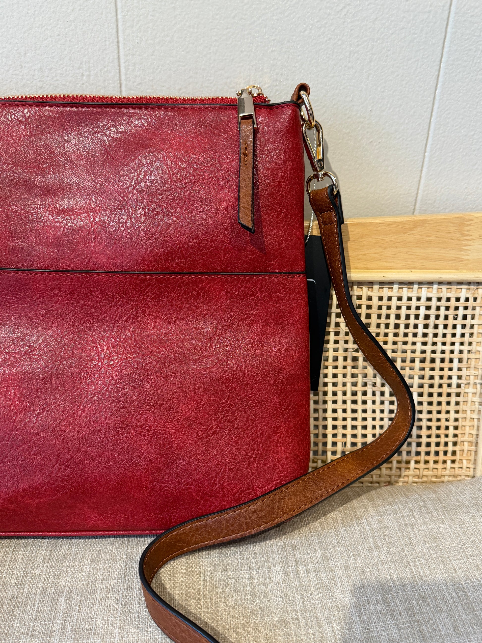A close-up of a red leather crossbody bag with a brown leather strap and zipper pull tab, resting on a textured beige fabric surface next to a woven cane basket.