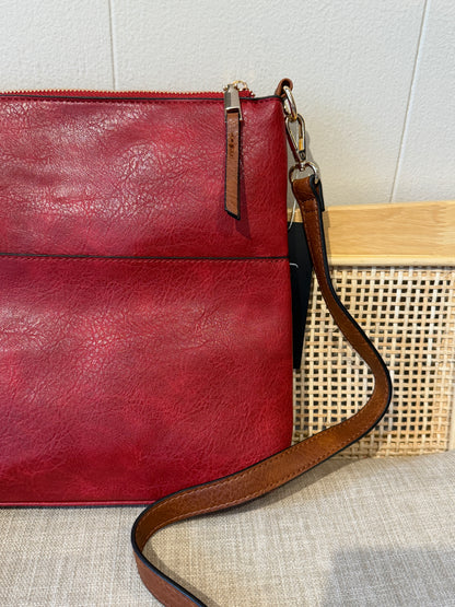 A close-up of a red leather crossbody bag with a brown leather strap and zipper pull tab, resting on a textured beige fabric surface next to a woven cane basket.