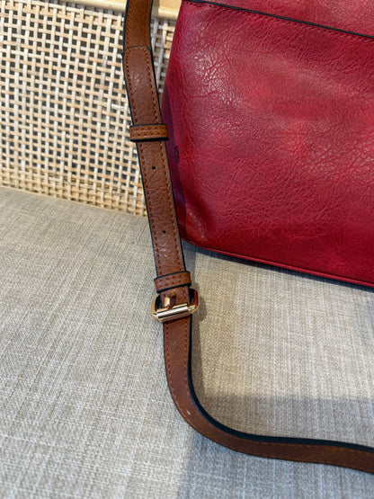A close-up of a red leather bag with a brown leather strap and gold-tone buckle, resting on a textured beige fabric surface.