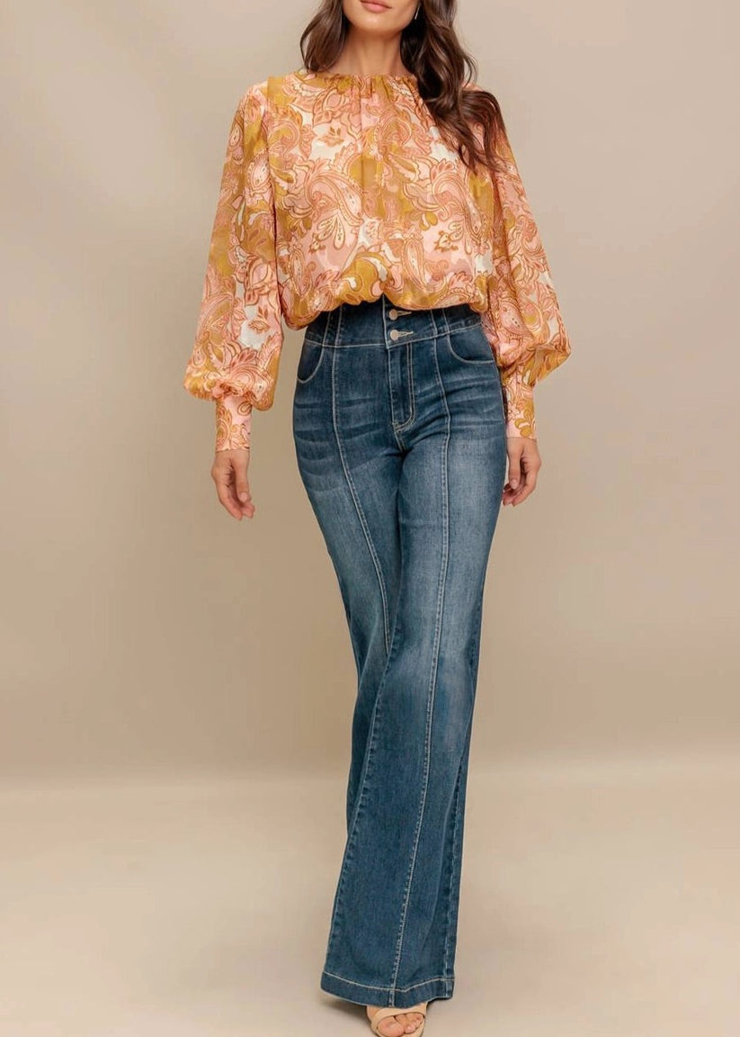 Woman wearing a camel, pink and ivory floral  paisley print blouse and jeans against a beige background.