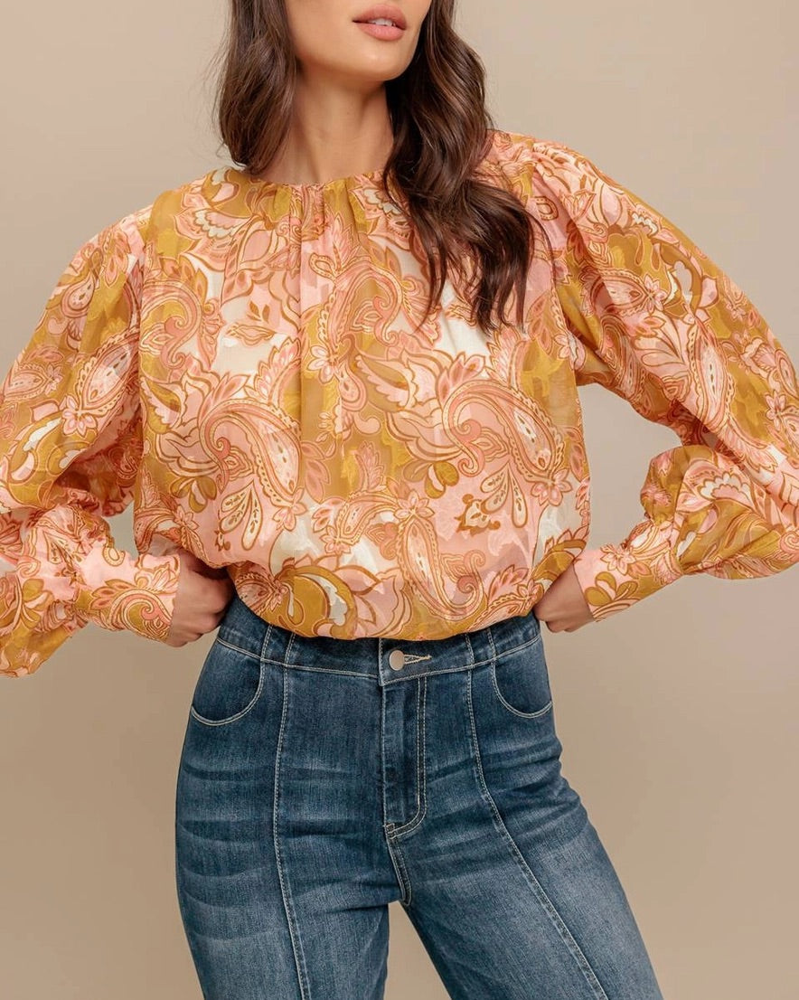 Woman wearing a camel, pink and ivory floral  paisley print blouse and jeans against a beige background. 