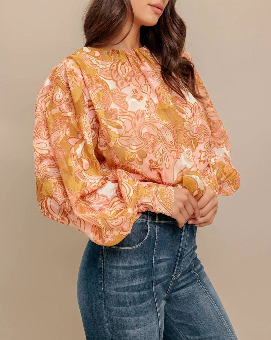 Woman wearing a camel, pink and ivory floral  paisley print blouse and jeans against a beige background. 