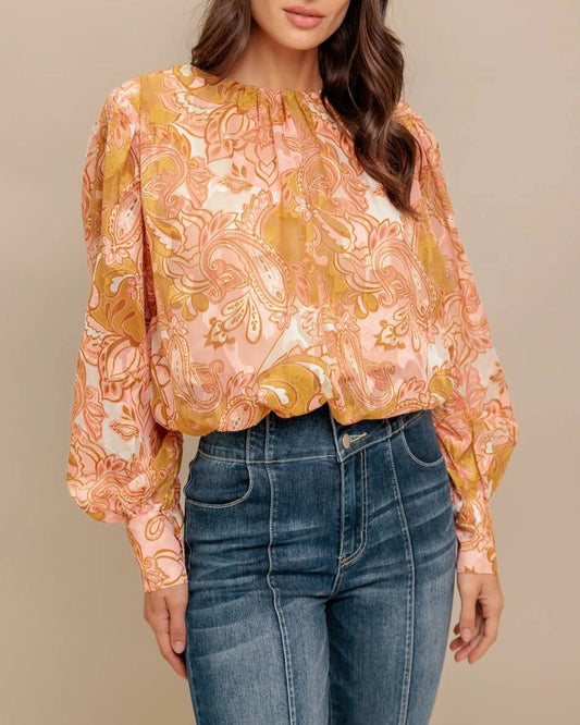 Woman wearing a camel, pink and ivory floral  paisley print blouse and jeans against a beige background.