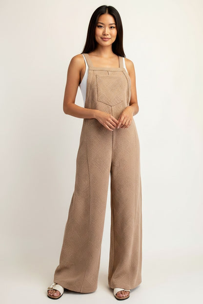 A woman modeling a camel wide-leg jumpsuit with a textured geometric pattern, styled over a white camisole and brown sandals against a plain light background.