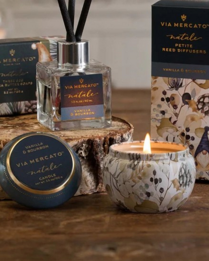 Via Mercato Natalie Vanilla & Bourbon scented products displayed on a rustic wooden surface, including a reed diffuser bottle with black reeds, a round candle tin, a lit floral-patterned candle jar, and branded boxes.