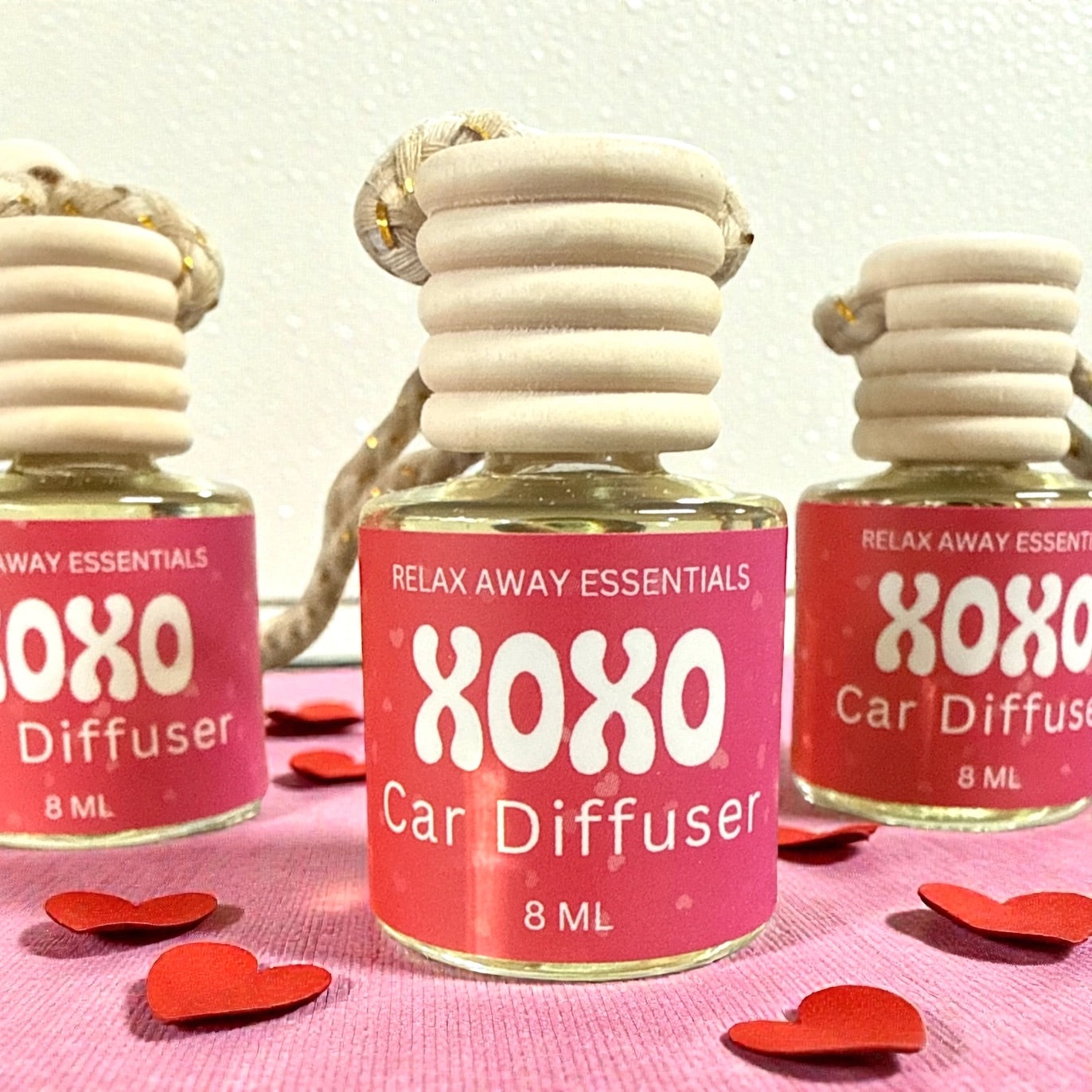 XOXO Car Diffuser bottles with pink labels on a pink background with red heart shapes.