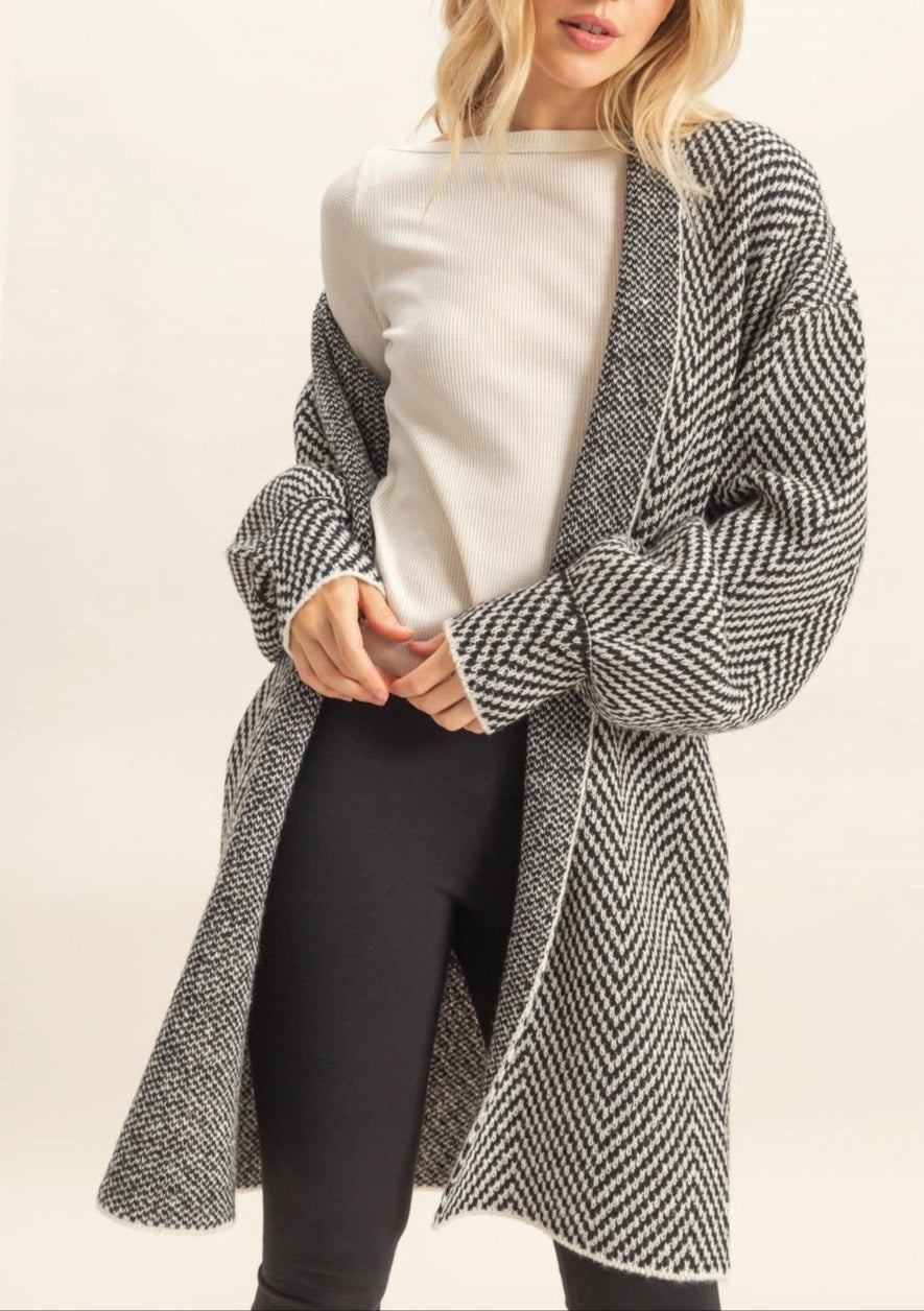 Woman wearing a long, B&W herringbone patterned cardigan over a white top and black pants on a beige background.