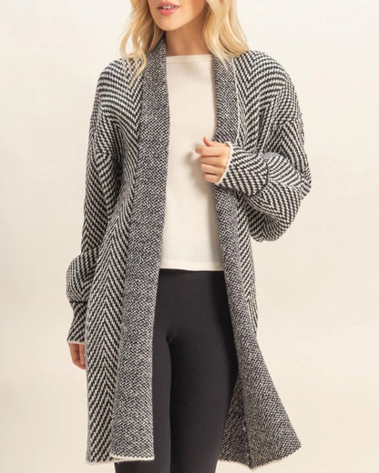 Woman wearing a long, B&W herringbone patterned cardigan over a white top and black pants on a beige background.