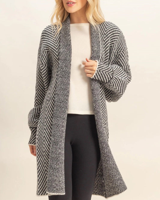 Woman wearing a long, B&W herringbone patterned cardigan over a white top and black pants on a beige background.