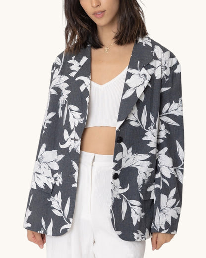 Woman wearing a charcoal floral patterned blazer over a white top and pants on a white background.