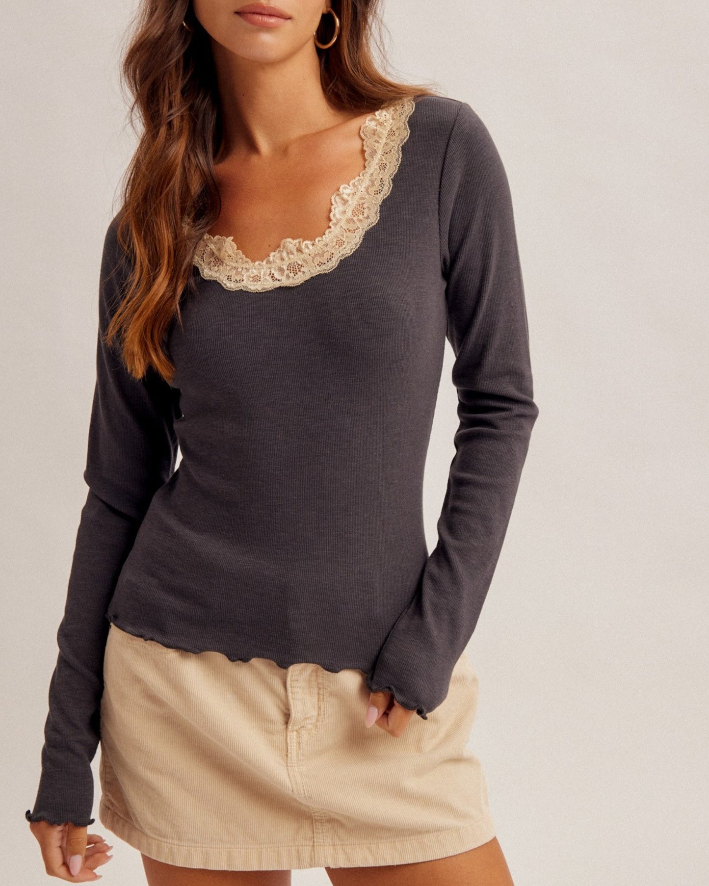 A woman wearing a dark charcoal gray long-sleeve top with a beige lace-trimmed scoop neckline, paired with a beige mini skirt, against a plain light gray studio background.