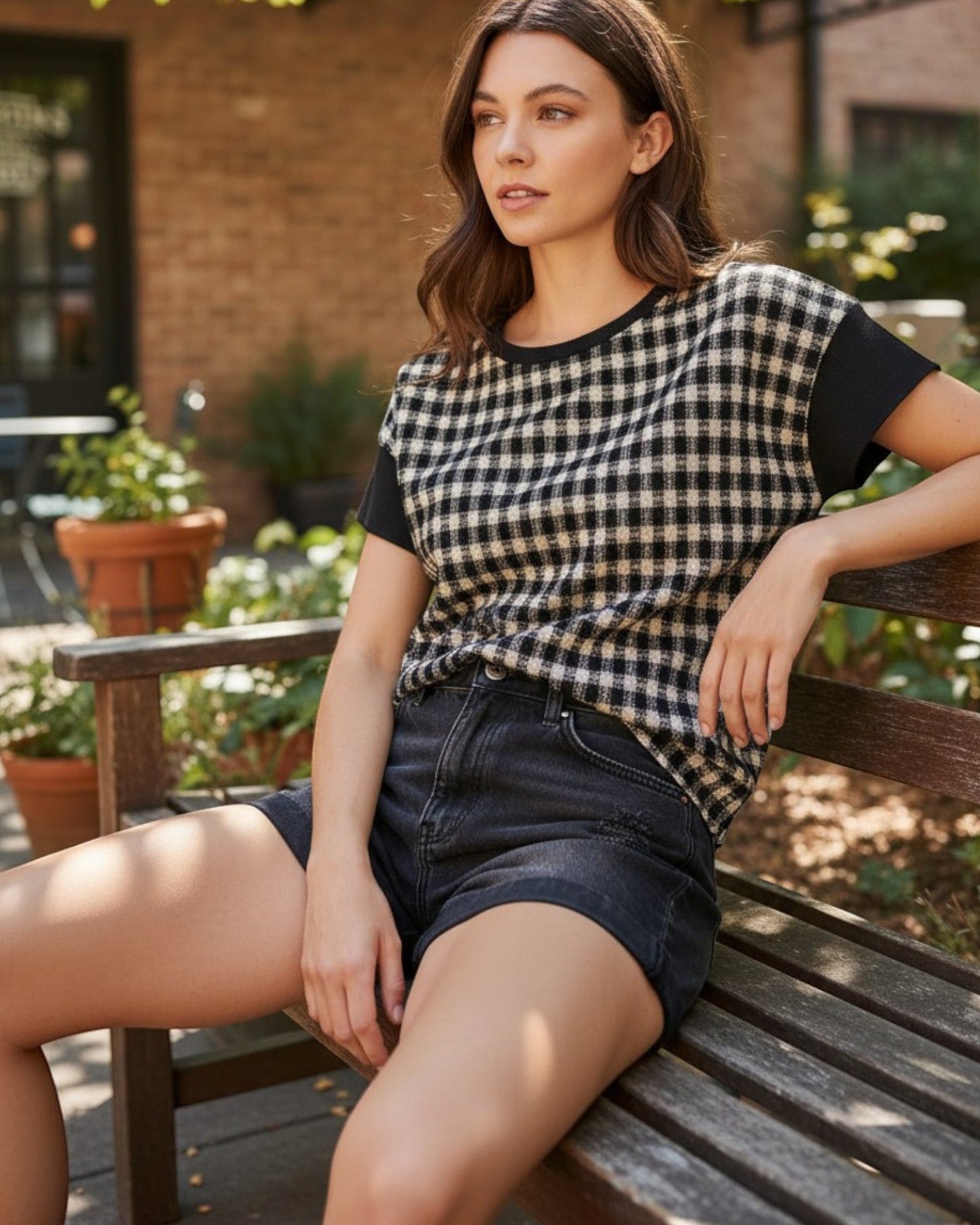 A woman sits on a wooden bench outdoors wearing a black and beige gingham short-sleeve top with solid black sleeves and dark denim shorts.
