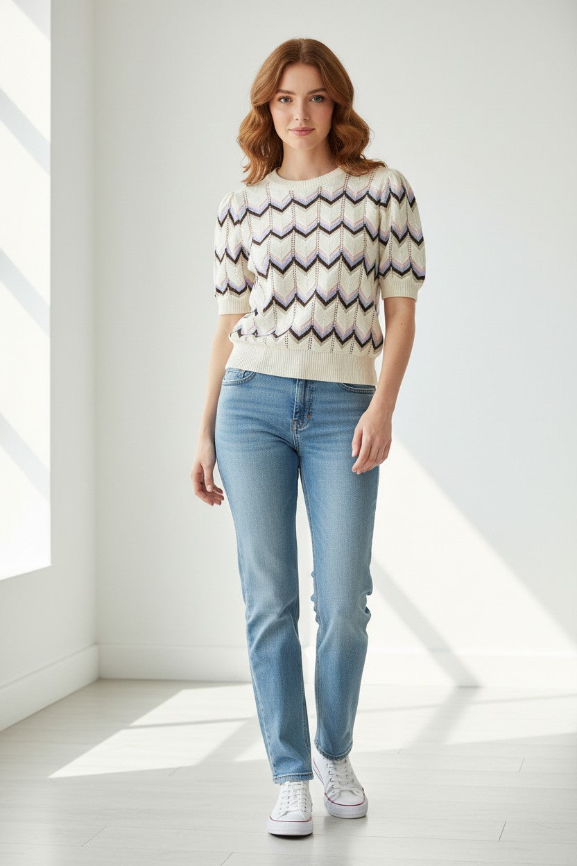 A woman stands in a bright white room wearing a cream knit sweater with a multi-color  stripe chevron pattern, light blue jeans, and white sneakers.