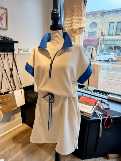 A cream-colored short-sleeve polo dress with blue collar and sleeve accents, displayed on a  mannequin inside a boutique. The dress features a half-zip neckline, a drawstring waist, and side pockets.