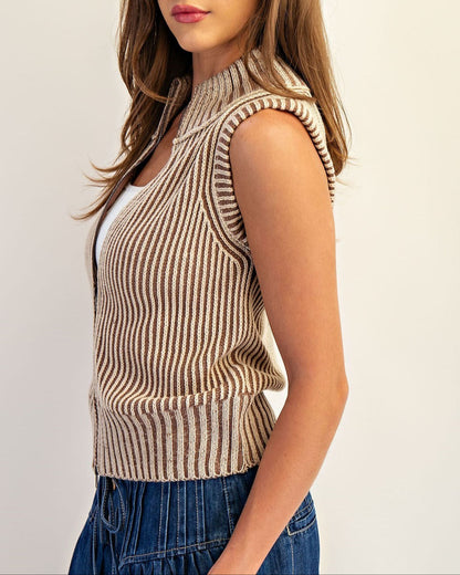 Woman wearing a tan striped ribbed vest over a white top and blue jeans on a plain background.