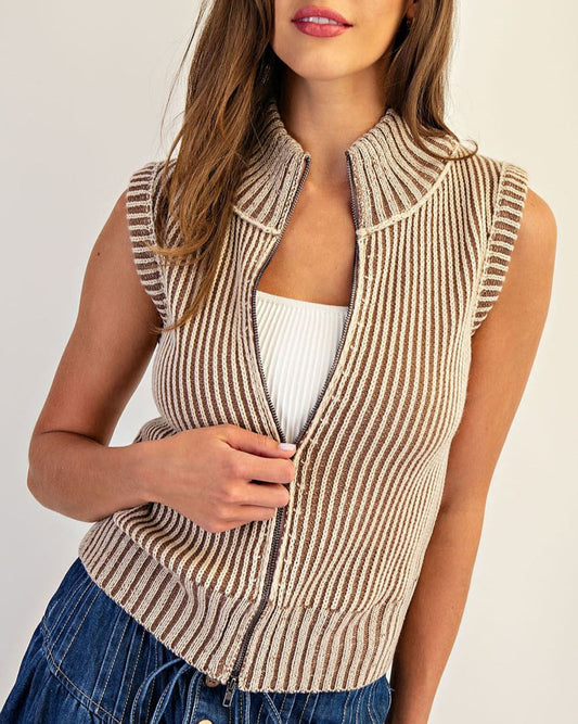 Woman wearing a tan striped ribbed vest over a white top and blue jeans on a plain background.