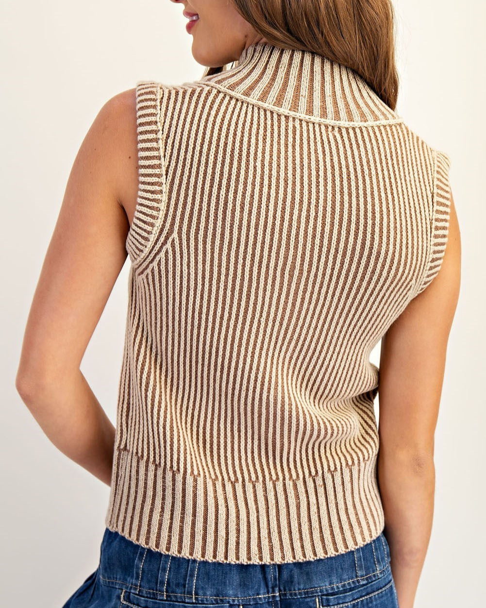 Woman wearing a tan striped ribbed vest over a white top and blue jeans on a plain background.