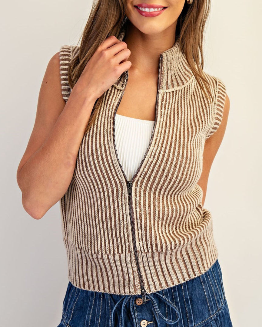 Woman wearing a tan striped ribbed vest over a white top and blue jeans on a plain background.