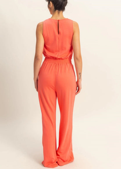 Woman wearing Sunset Hour Sleeveless Jumpsuit in Coral with cinched waist and high scoop neckline. 