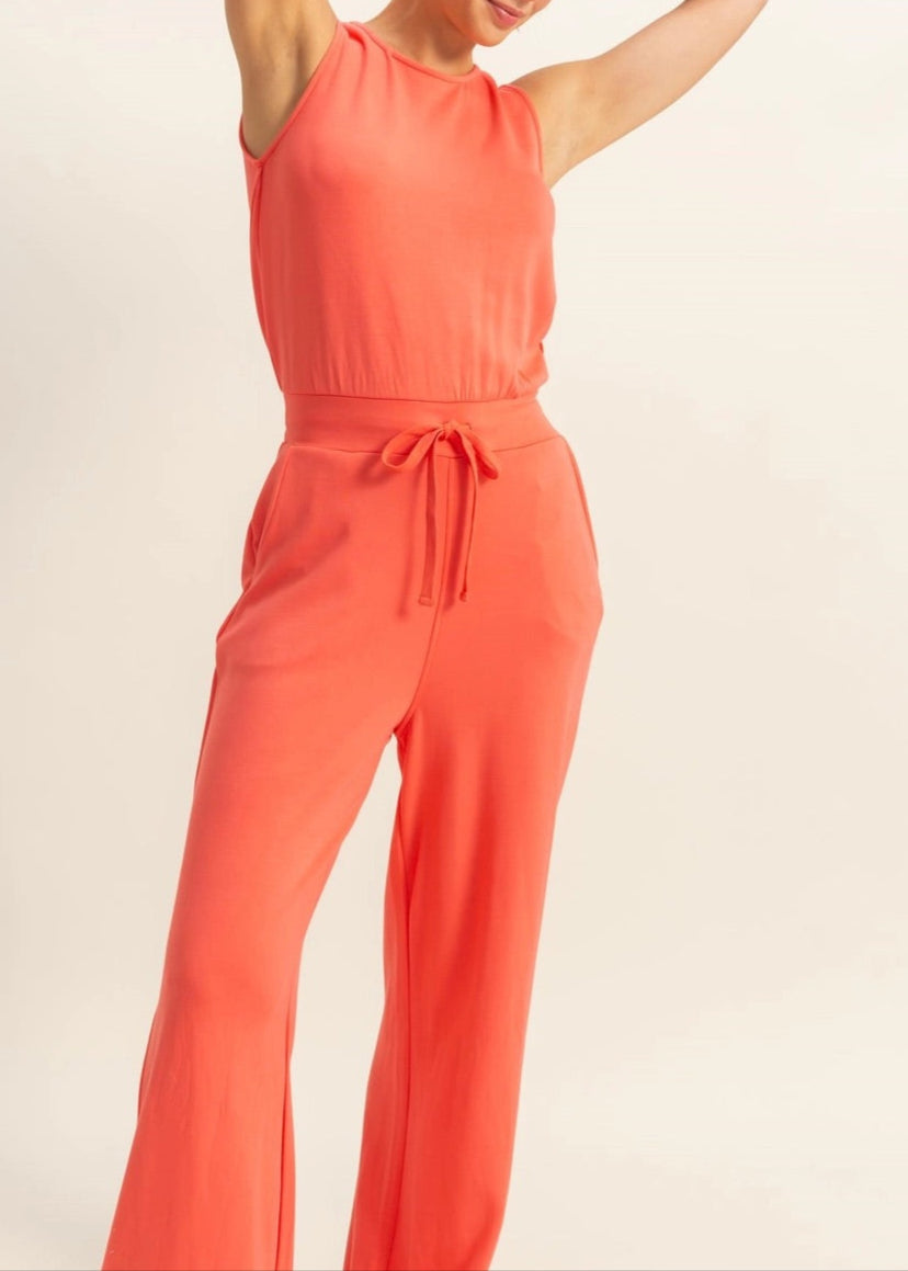 Woman wearing Sunset Hour Sleeveless Jumpsuit in Coral with cinched waist and high scoop neckline. 