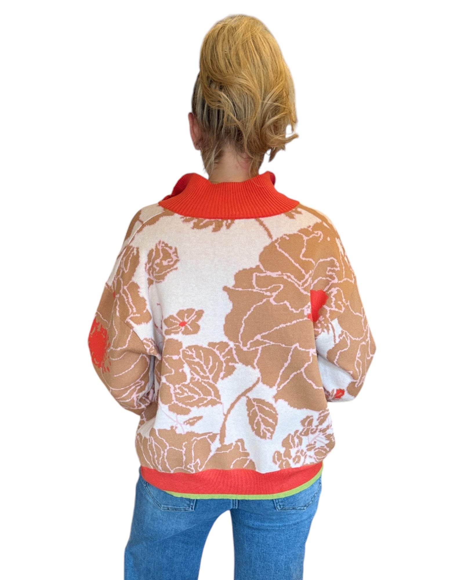 Woman wearing a coral, tan and cream half zip floral pattern sweater and blue jeans. 