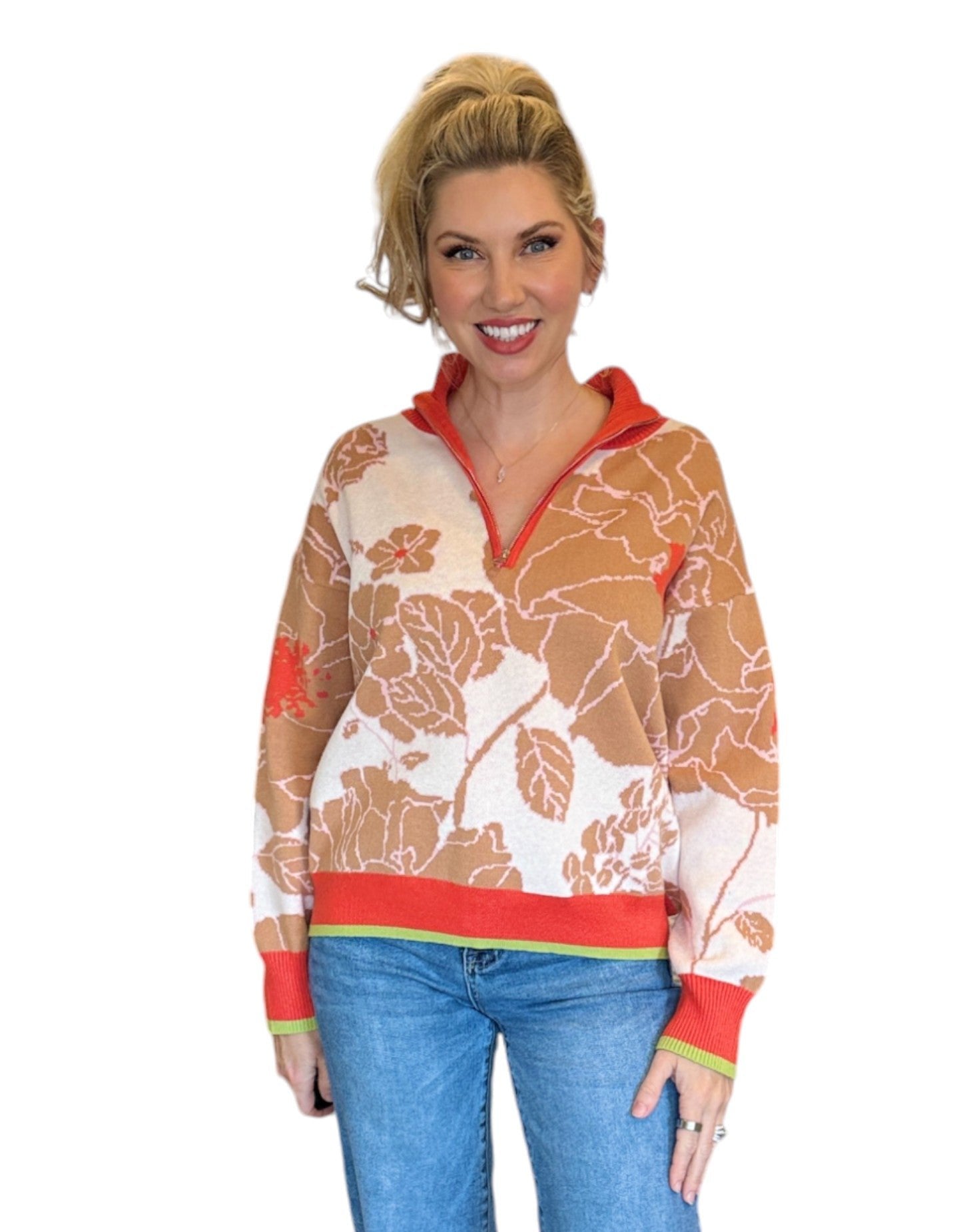 Woman wearing a coral, tan and cream half zip floral pattern sweater and blue jeans. 