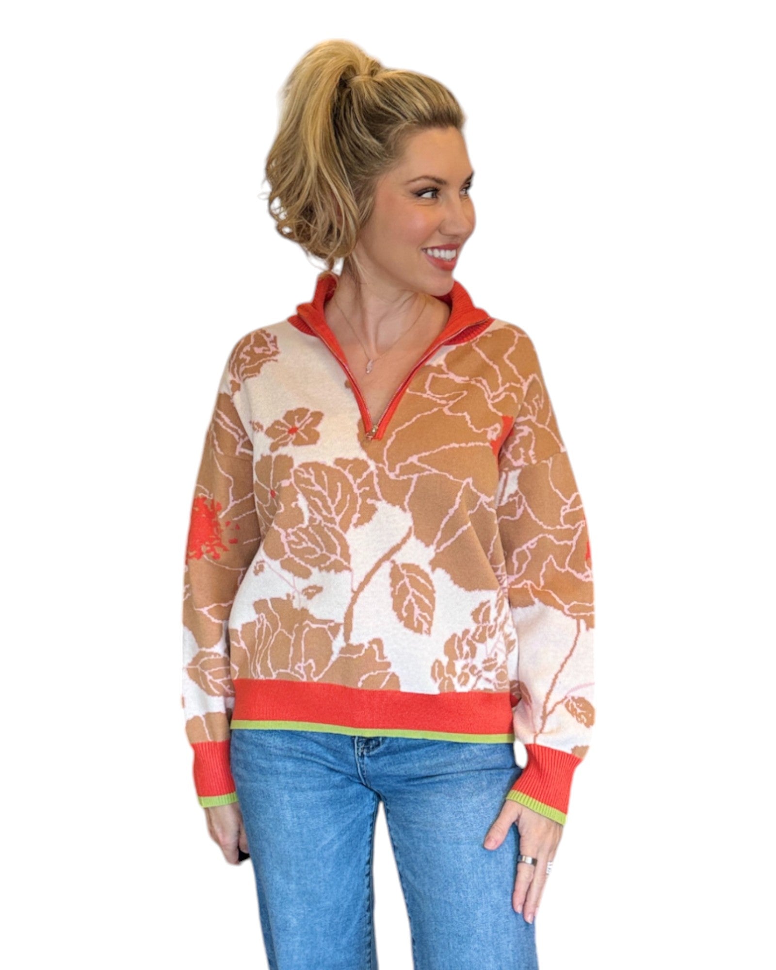 Woman wearing a coral, tan and cream half zip floral pattern sweater and blue jeans. 