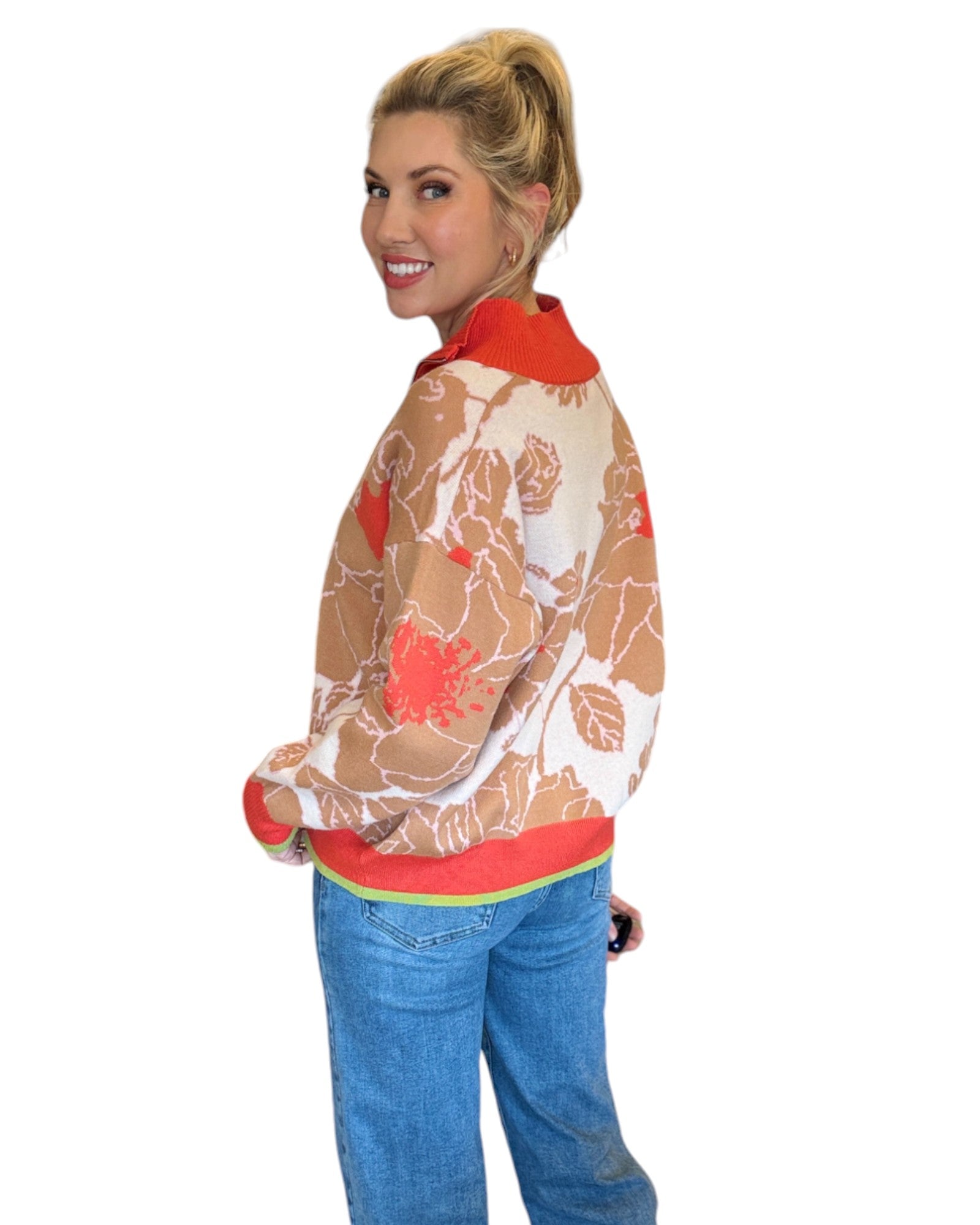 Woman wearing a coral, tan and cream half zip floral pattern sweater and blue jeans. 