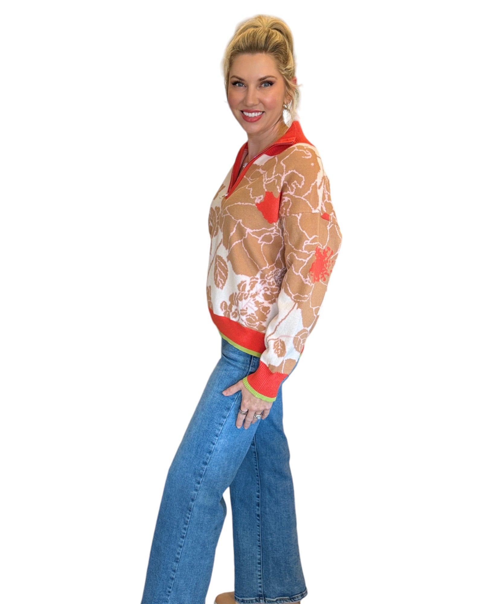 Woman wearing a coral, tan and cream half zip floral pattern sweater and blue jeans. 