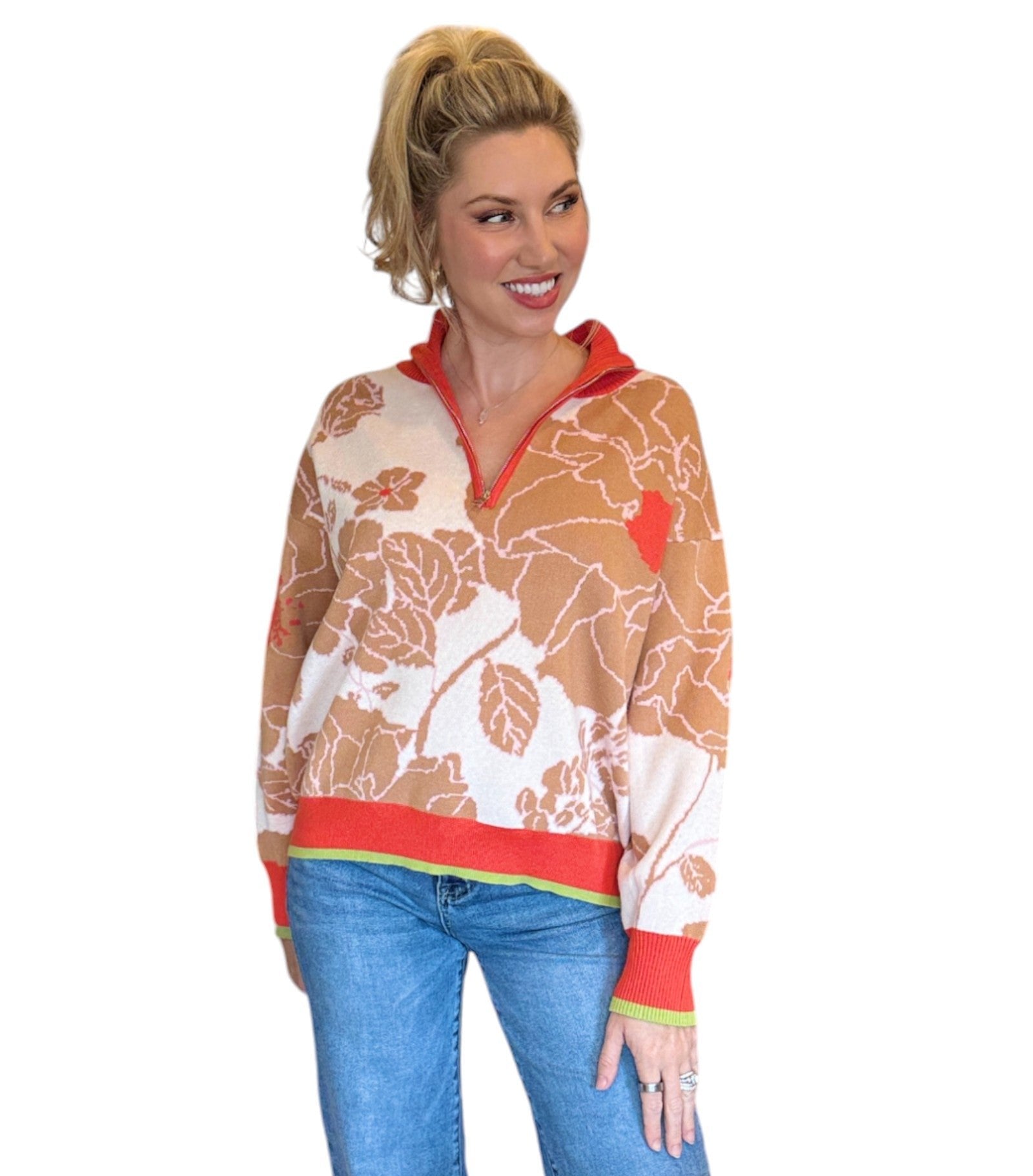 Woman wearing a coral, tan and cream half zip floral pattern sweater and blue jeans. 