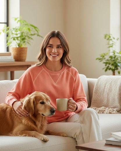 Woman in coral wash sweatshirt sitting on a couch with a dog and holding a mug in a cozy living room.
