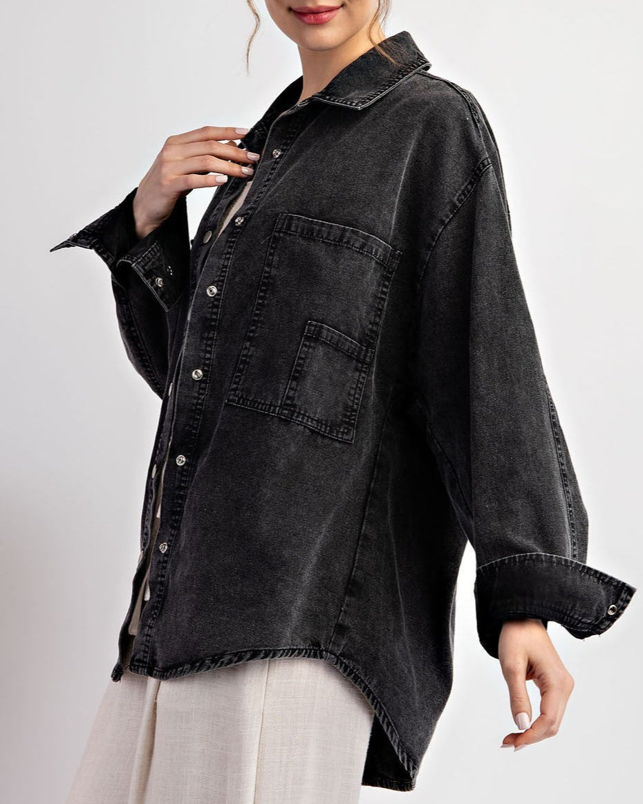 Woman wearing a black denim jacket / shacket over a beige top and white pants on a light gray background.