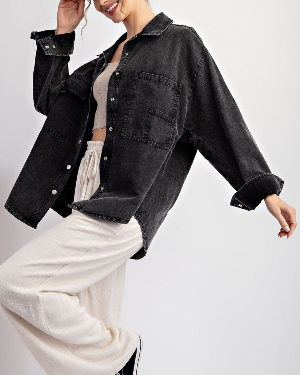Woman wearing a black denim jacket / shacket over a beige top and white pants on a light gray background.