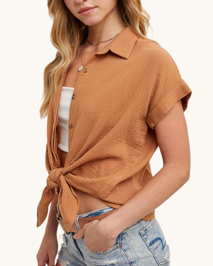 Woman wearing a brown tied shirt over a white top and denim shorts on a white background.