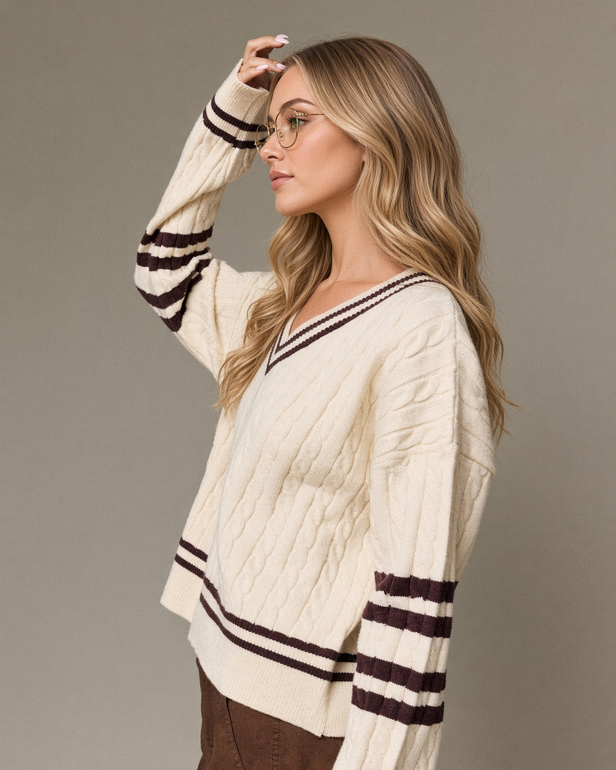Side view model wearing a cream cable knit V-neck sweater with brown striped neckline, sleeves and hem.