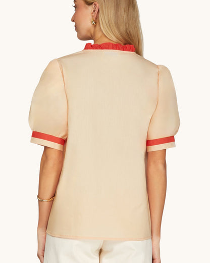 A woman shown from the back wearing a light beige short-sleeve blouse with a red ruffled collar and red stripe accents at the sleeve cuffs, paired with white pants against a light beige studio background.