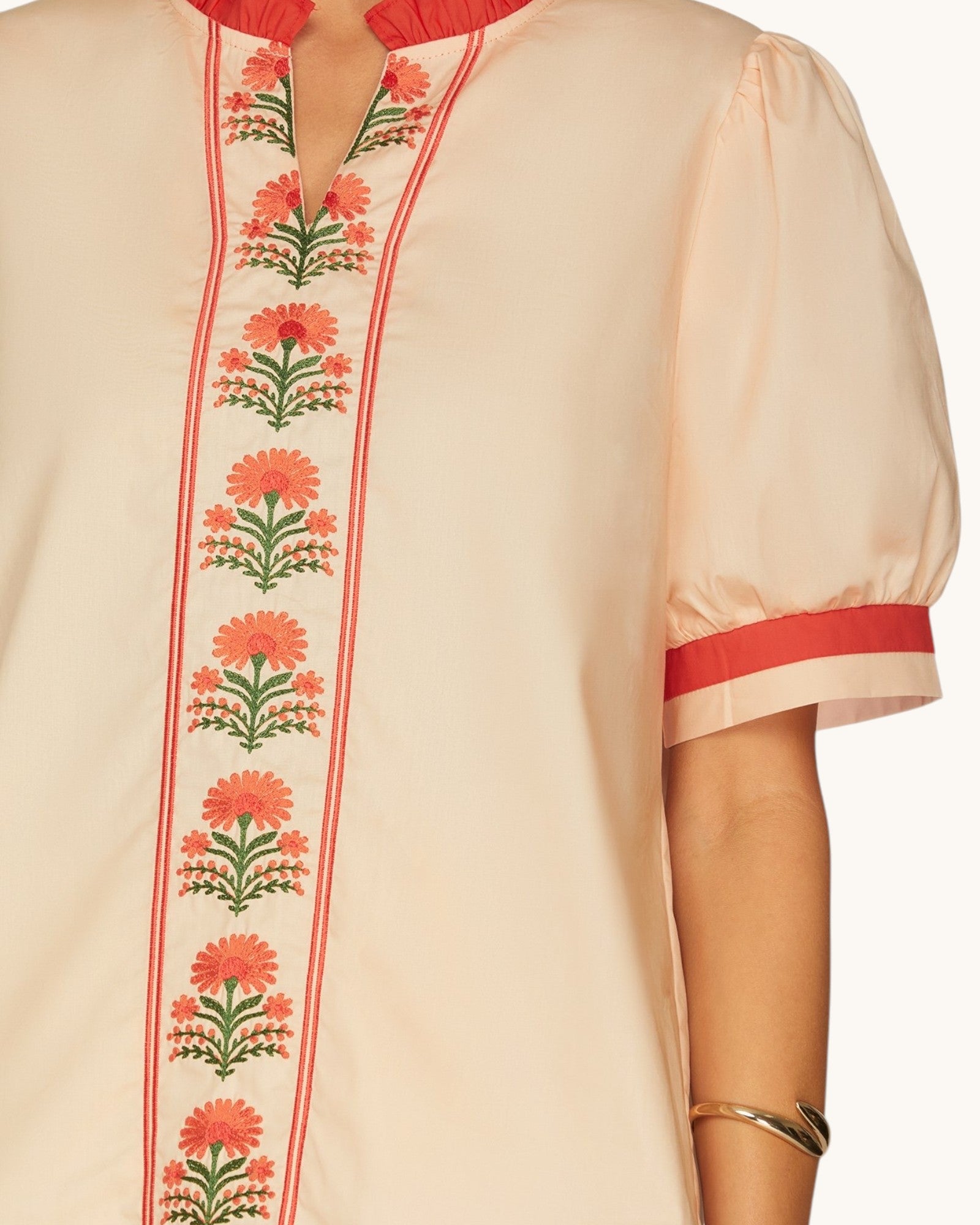 A close-up of a peach-colored tunic top with red and green floral embroidery down the front placket and red trim on the collar and sleeve cuffs, worn by a model.