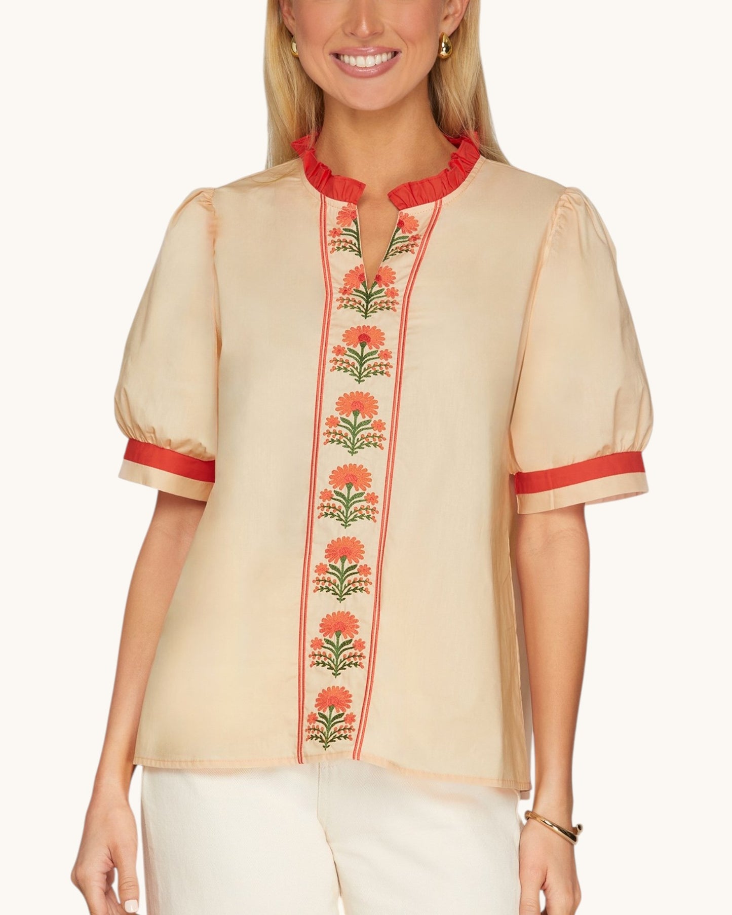 A woman wearing a peach short-sleeve blouse with red ruffled trim at the collar and cuffs, and a vertical floral embroidered panel down the front.