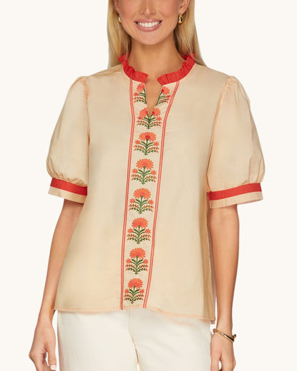 A woman wearing a peach short-sleeve blouse with red ruffled trim at the collar and cuffs, and a vertical floral embroidered panel down the front.