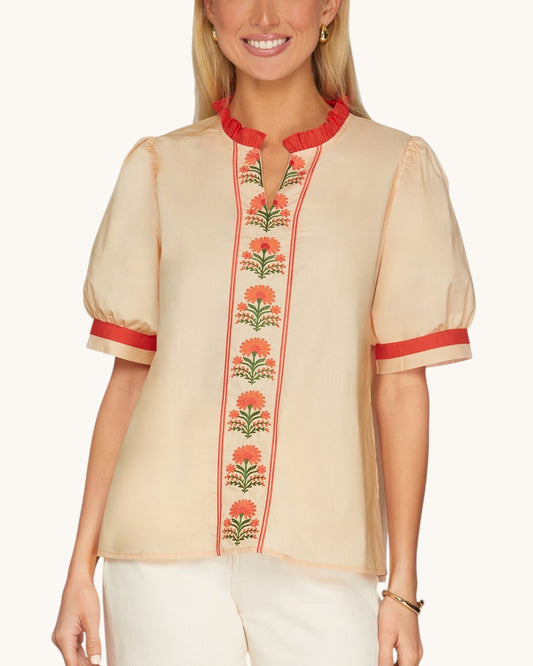 A woman wearing a peach short-sleeve blouse with red ruffled trim at the collar and cuffs, and a vertical floral embroidered panel down the front.