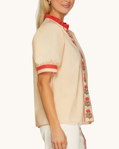 A side view of a woman wearing a light peach short-sleeve blouse with red ruffled trim at the collar and cuffs, and red floral embroidery down the front placket, paired with white pants against a light cream studio background.
