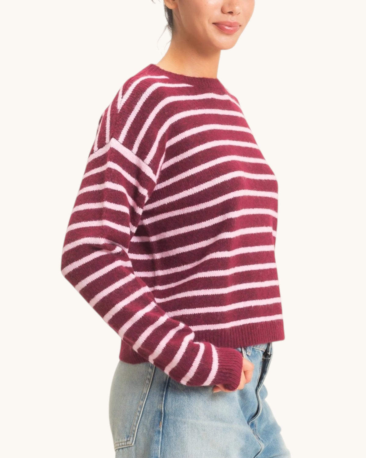 A woman wearing a maroon and light pink striped knit sweater with light blue jeans, shown from a side profile against a plain off-white background.
