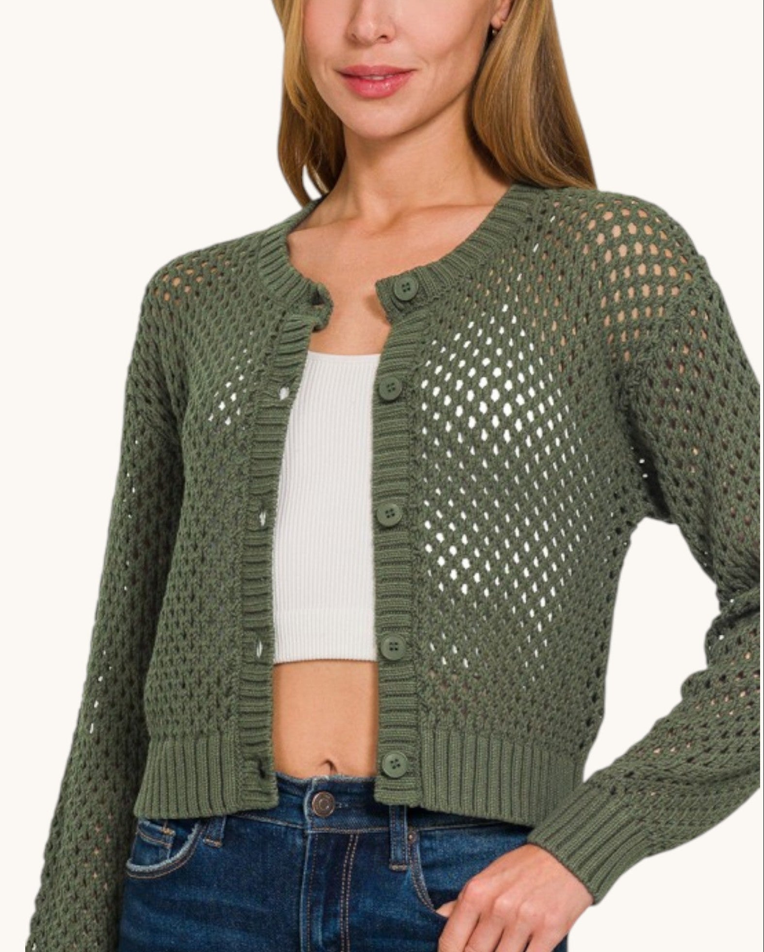 A woman wearing an olive green open-knit cropped cardigan with button closures over a white ribbed crop top and blue jeans, shown against a plain light background.