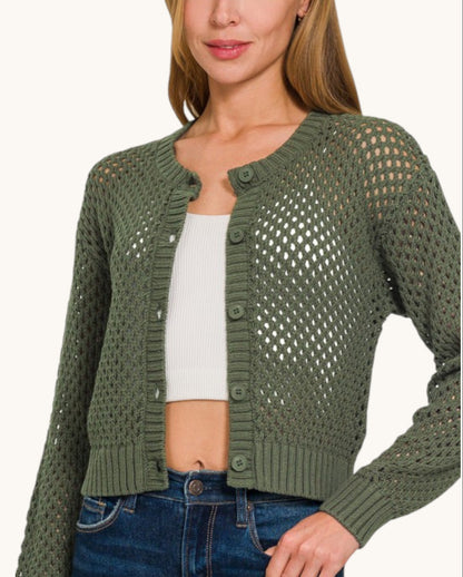 A woman wearing an olive green open-knit cropped cardigan with button closures over a white ribbed crop top and blue jeans, shown against a plain light background.
